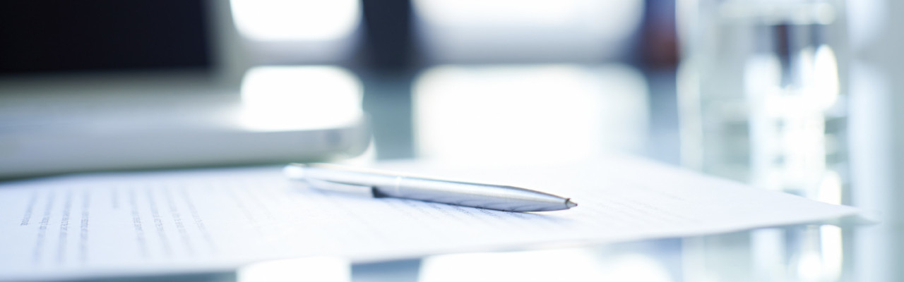 Silver pen and business contract situated on office desk, people in the background